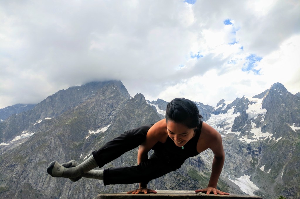 A woman dressed in all black doing an arm balancing yoga pose, eight angle pose, in an outdoor, alpine, mountainous setting with glaciers