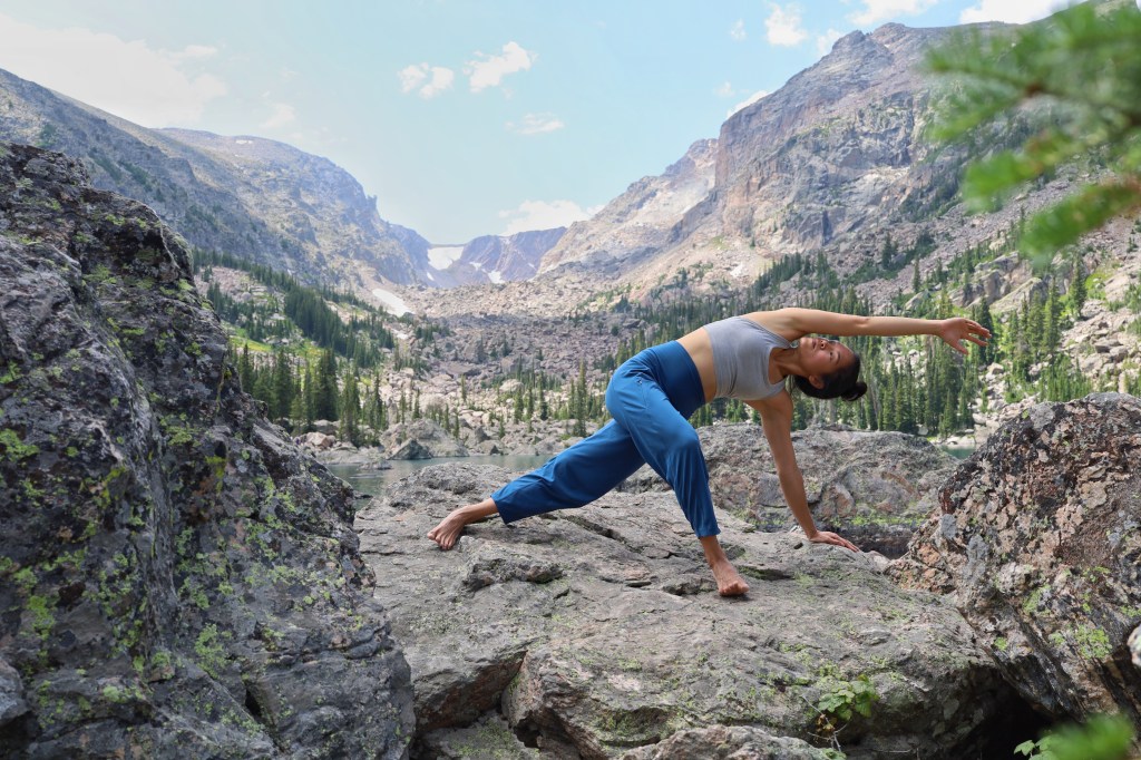 A woman doing outdoor yoga, in the pose horizon lunge, with beautiful mountains, pine trees, and an alpine lake behind her