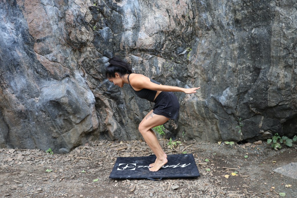 A woman dressed in all black doing yoga in the pose Drinking Bird, a variation of chair pose, in front of an outdoor rock climbing boulder