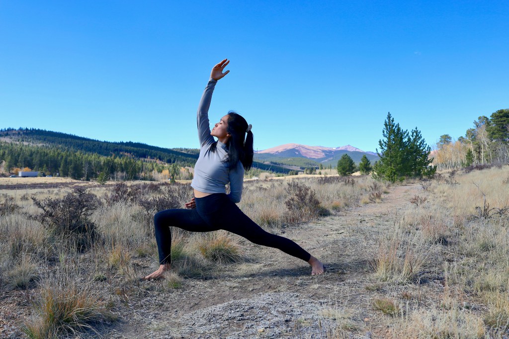 A woman doing outdoor yoga, in the pose reverse warrior 2, with mountains and trees behind her