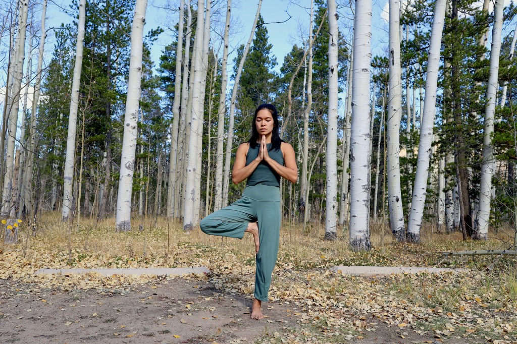 A woman dressed in green doing outdoor yoga, standing in Tree Pose, with aspen and pine trees behind her