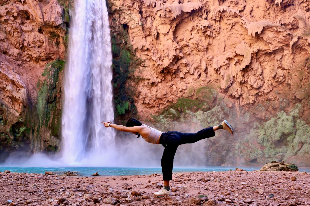 A woman standing in the yoga pose Warrior 3, in front of a turquoise blue waterfall and sandstone cliff.