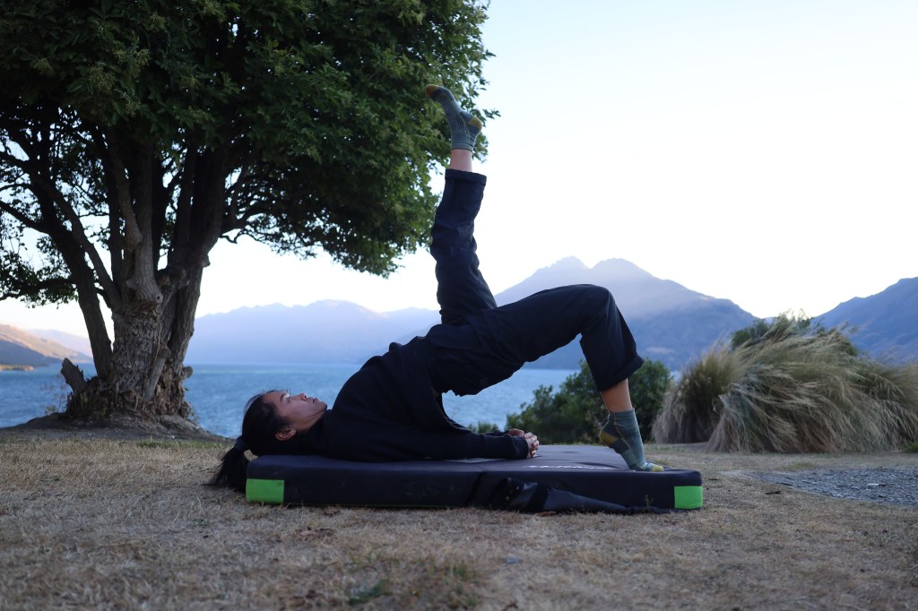 A woman in a variation of the yoga pose bridge pose in an outdoor mountain seascape setting at dusk